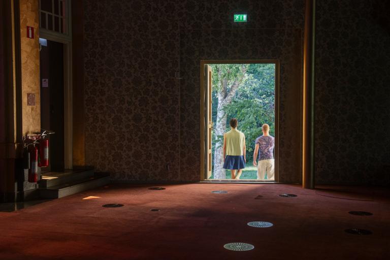 Två personer går ut från en mörkt rum ut i en parkliknande miljö.