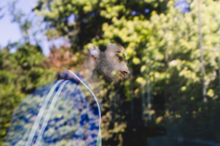 Dansaren Facundo står vid ett stort fönster. Han är klädd i en blå träningsjacka med vita ränder. Han har kort mörkt hår och kort skägg. Facundo täcks nästan helt av reflektionen i fönstret av stora grönskande träd.