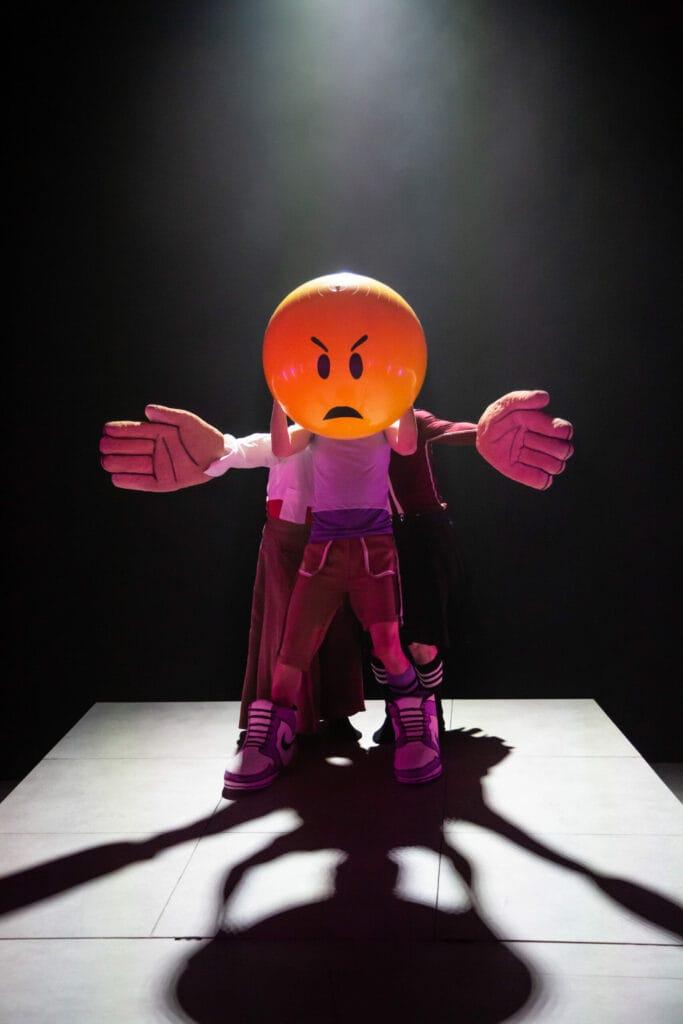 En dansare i ett stort emojuformat huvud med argt uttryck och stora dockarmar.