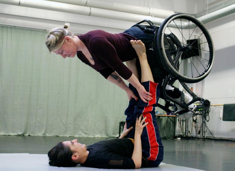 En dansare ligger på golvet med ben sträckta mot taket. Balanserande på de sträckta benen ligger en annan dansare. 