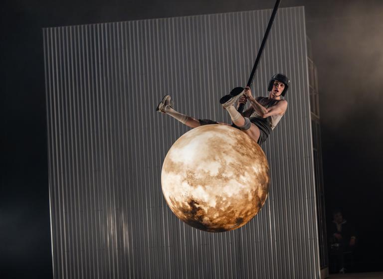 Dansaren Carl sitter och gungar på en rivningskula som ser ut som en fullmåne. Kulan svingar genom luften och CArl i discobollshjälm sträcker ut benen för att ta mer fart i Skånes DAansteaters jubileumsföreställning Wing Ding av koreografen Lidia Wos.