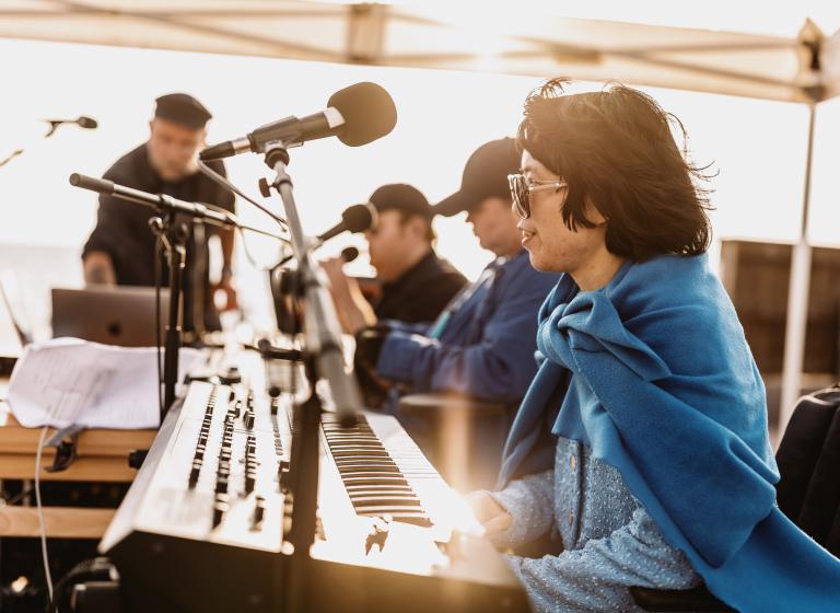 Musiker sitter i solljuset klädda i blå nyanser vid ett bord med keyboards och mikrofoner i Projekt Viaductus föreställning Blå timme.