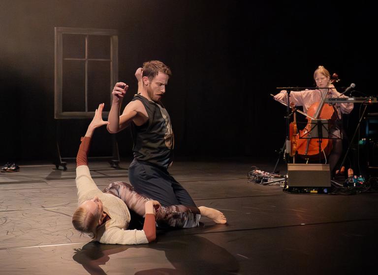 Dansaren Jure sitter på dansaren Anette som ligger på golvet. I bakgrunden spelar Emma Augustsson cello. 