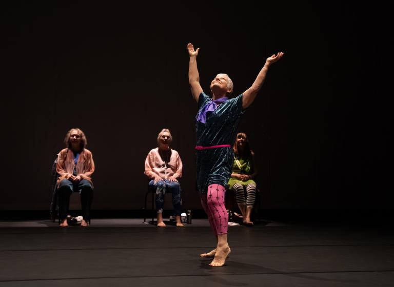 En gästdansare över 60 år klädd i blå tunika och rosa tightssträcker armarna uppåt och vänder sitt leende ansikte uppåt i dansföreställningen Jag minns mig själv.