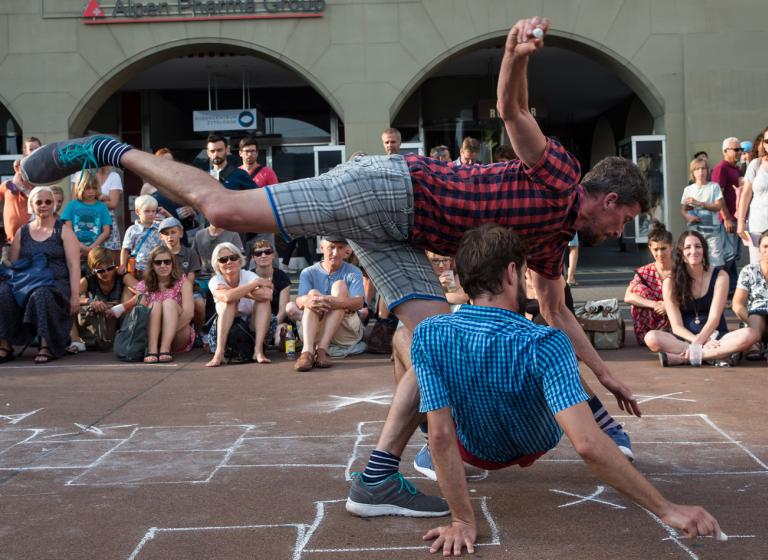 Två dansare hoppar hage framför en folksamling. Ena dansaen sträcker sig med ett ben över den andra dansaren som balanserar på händer och fötter på rygg.