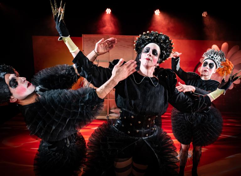 Tre dansare med svart smink runt ögonen, klädda i svarta bulliga kläder står tillsammans. Två av dem fixar med den tredje som ställt upp sig mellan dom.