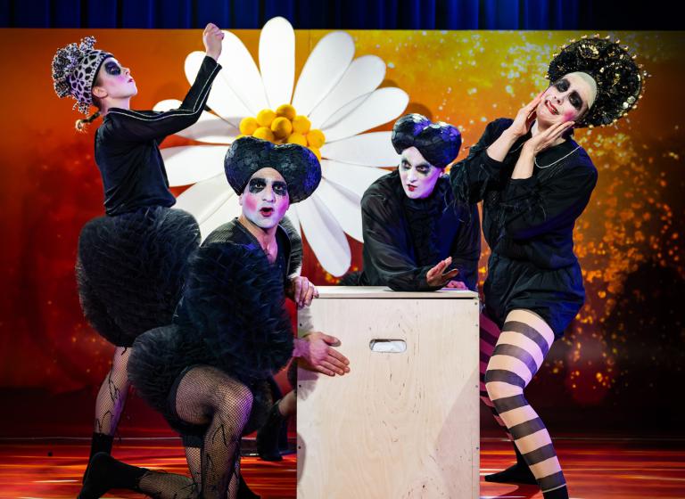 Fyra dansare med bulliga svarta byxor och ovanliga hattar står vid ett vitt litet skåp och en enorm prästkrage och ser lite oroliga ut.