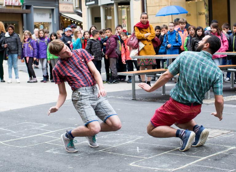 Två dansare i shorts och rutiga skjortor balanserar på tå mittemot varandra på en lekhage på en gata.