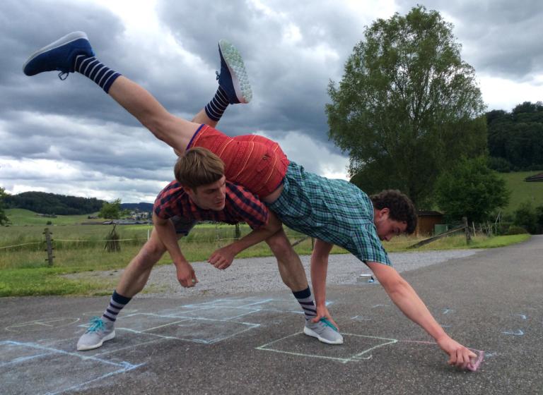 En dansar balanserar på en annan dansares rygg medan han ritar upp en hopphage på marken.