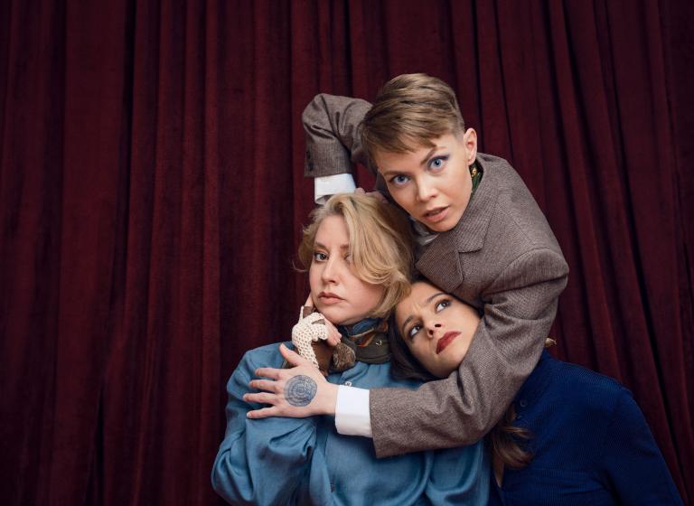 Dansaren Emma håller om dansarna Madeleine och Marion. Madeleine ser fundersam ut och Marion tittar misstänksamt på Emma.