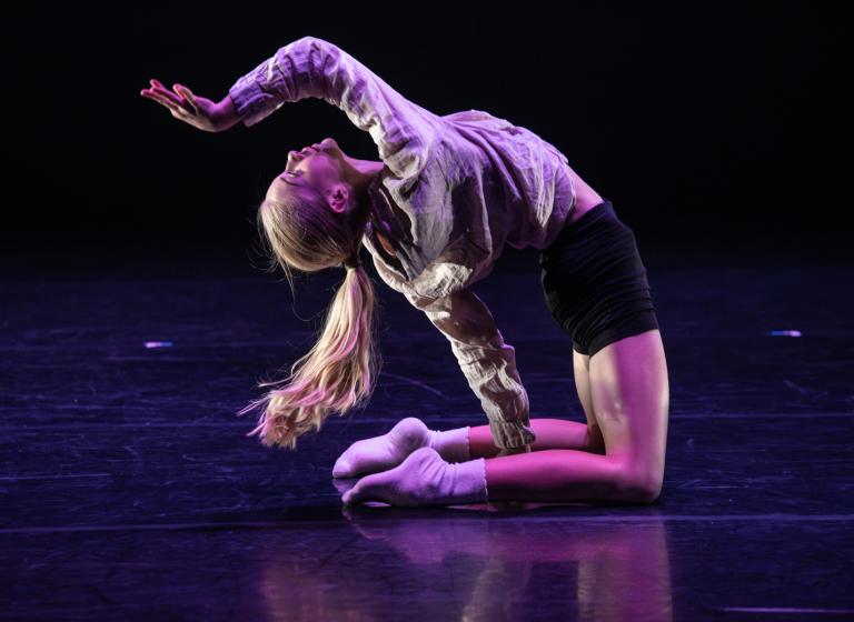 En ung dansare på knä böjer sig bakåt med en arm böjd ovanför sitt ansikte.