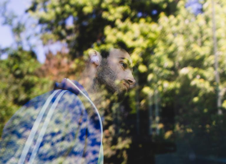 Dansaren Facundo står vid ett stort fönster. Han är klädd i en blå träningsjacka med vita ränder. Han har kort mörkt hår och kort skägg. Facundo täcks nästan helt av reflektionen i fönstret av stora grönskande träd.