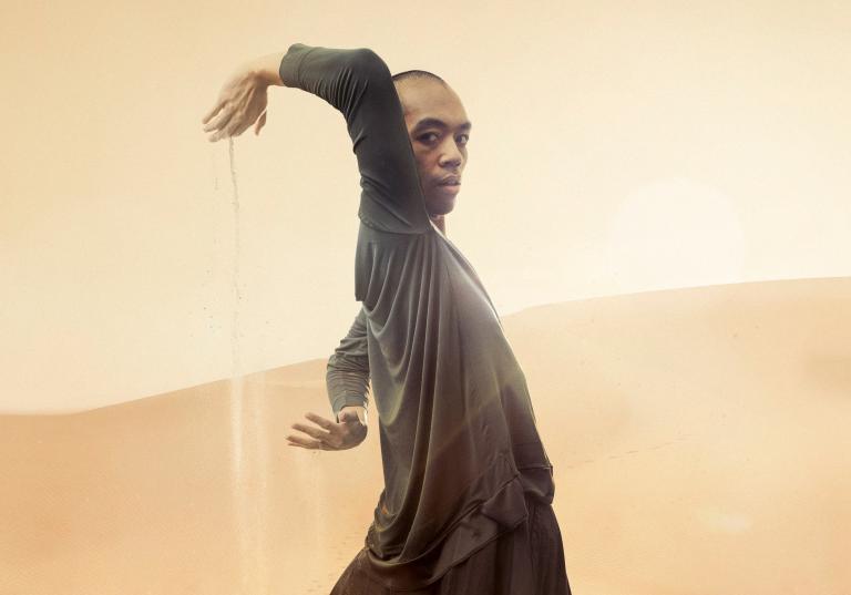 Dansaren Micahel står i ett gyllene ökenlandskap med armarna böjda bakom sig i en cirkel och sand strilande från sina händer.