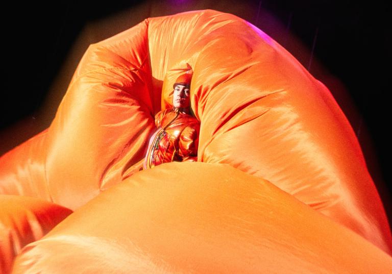 En dansare i orange keps och glansig jacka är omgiven av ett molnliknande orange tyg.