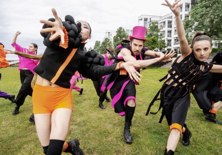 Dansare i svarta kläder med mycket orange och rosa detaljer dansar mot kameran.
