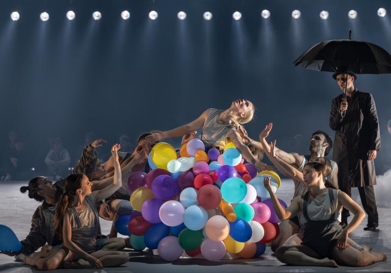 Dansaren Matilda kläd i en stor kjol täckt av ballonger i regnbågens alla färger står omringad av dansare som sitter på golvet runt henne. Bakom dem står en man i mörk kostym och paraply. En del av Skånes Dansteaters jubileumsföreställning Wing Ding.