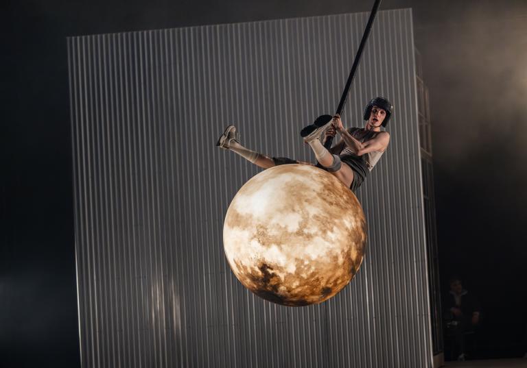 Dansaren Carl sitter och gungar på en rivningskula som ser ut som en fullmåne.  Kulan svingar genom luften och CArl i discobollshjälm sträcker ut benen för att ta mer fart i Skånes DAansteaters jubileumsföreställning Wing Ding av koreografen Lidia Wos.