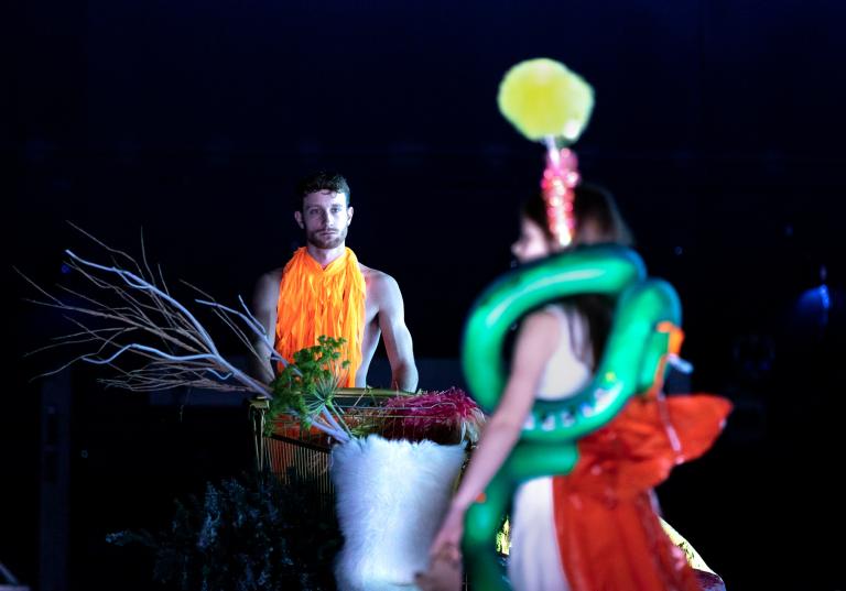 Dansaren Jure kommer gående i bakgrunden med en kundvagn fylld med kvistar och grenar. Han går mot en ung flicka som står i förgrunden klädd i ballonger i Skånes Dansteaters dansföreställning Out of the Blue.
