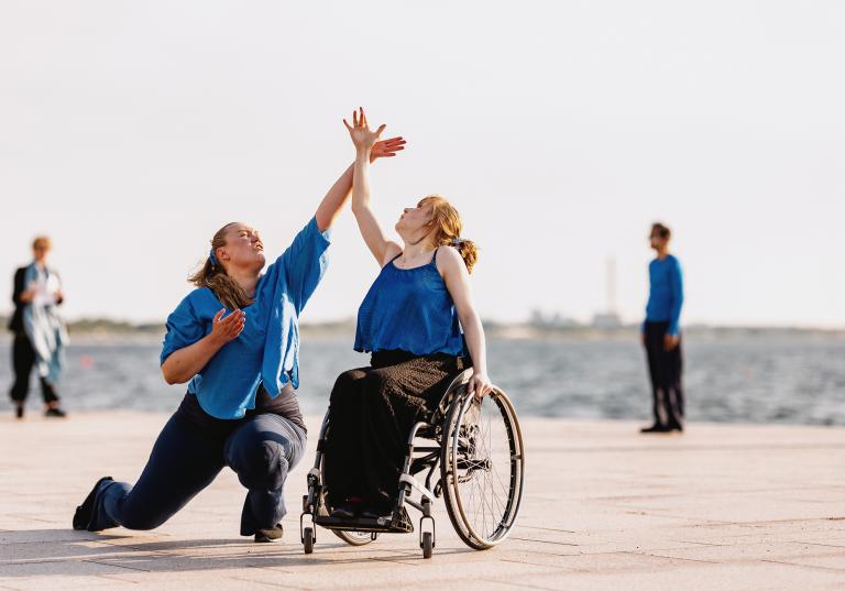 En dansare klädd i lång svart kjol och blått löst linne sitter i rullstol och sträcker upp sin arm mot himlen. Bredvid henne sträcker en annan dansare upp sin hand så att deras handleder möts i Projekt Viaductus föreställning Blå timme.