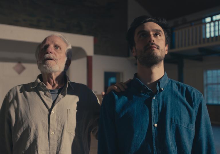 Dansarna Kit Brown i blå skjorta och Niels Simonsen i beige skjorta tittar snett upp mot taket i ett stort rum i Skånes Dansteaters dansfilm Eller så talar vi inte.