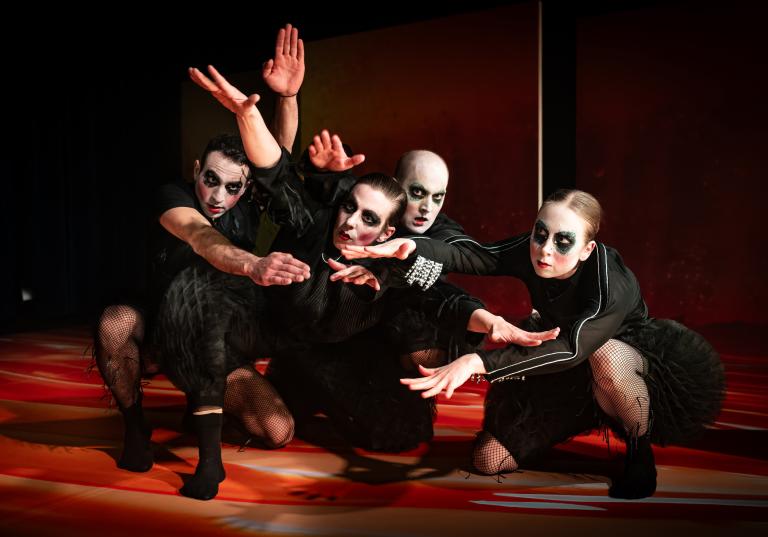 Fyra dansare hukar tillsammans på golvet och sträcker sina armar framför varandra. De har svarta bulliga kläder och svart smink runt ögonen i dansföreställningen Honey Honey.