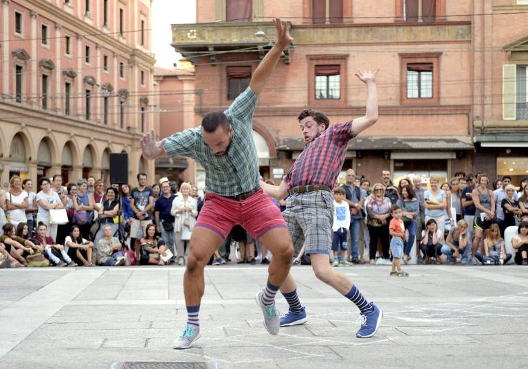 Två dansare i rutiga skjortor och shorts dansar yvigt utomhus på ett torg omgivna av männsikor.