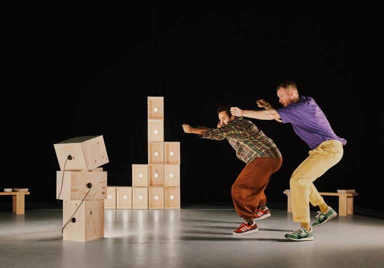 Dansarna Yared och Viktor dansar bredvid varandra bland omålade trälådor med lampor och sladdar i. De siktar tillsammans som om de hade pilbågar mot något vi inte ser.