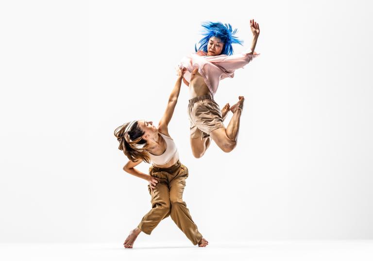 Dansaren Hanna hukar och sträcker sig upp mot dansaren Kat som hoppar högt upp i luften.