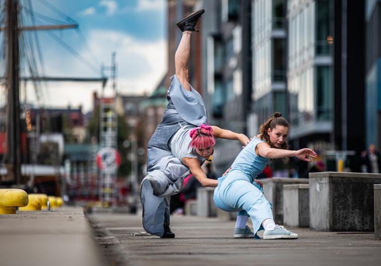 Dansarna Kat och Jeanne klädda i lösa ljusblå kläder dansar tillsammans vid segelbåtar vid kajkanten i Malmö hamn.