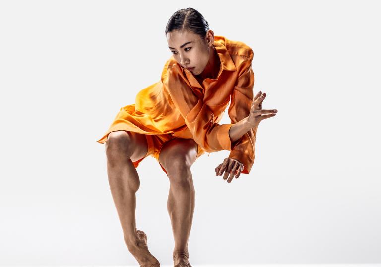 Dansaren Jing Yi Wang klädd i orange shorts och skjorta med svart uppsatt hår dansar med böjda knän och sträcker ut en hand.