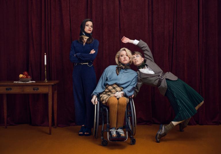 Dansarna Marion, Madeleine och Emma står framför en mörkröd ridå. Madeleine och Emma lutar sig mot varandra och i brakgrunden står Marion vid ett bord med ett tänt ljus och en skål med äpplen. 