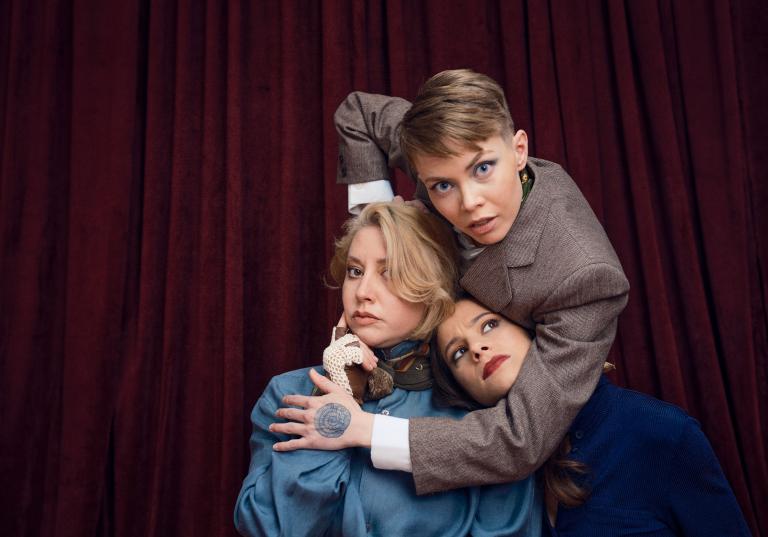 Dansaren Emma håller om dansarna Madeleine och Marion. Madeleine ser fundersam ut och Marion tittar misstänksamt på Emma.