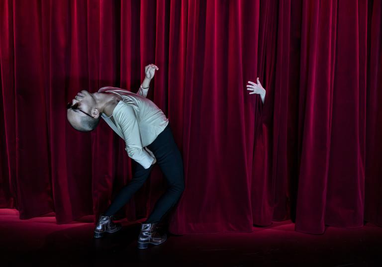 Dansaren Michael klädd i mörka byxor och en ljus draperad blus och solglasögon böjer sig kraftigt baklänges. Framför honom sticker en hand ut genom en mörkröd ridå.
