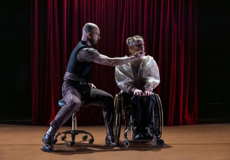 Dansaren Tomáš sitter på en pall på hjul. Han vänder sig och sträcker ut en hand mot dansaren Madeleines kind. Madeleine sitter i rullstol bredvd honom. 