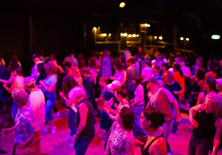 En mycket stor grupp människor dansar på ett stort dansgolv i ett rosa-lila ljus.