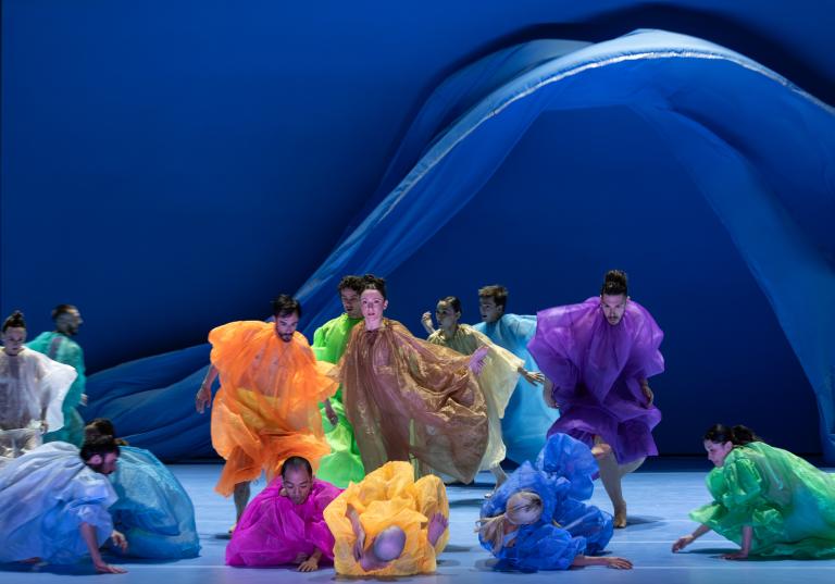 Dansare i skira overaller i klara färger dansar medan stora blå skynken faller bakom dem i Skånes Dansteaters dansföreställning Out of the Blue.