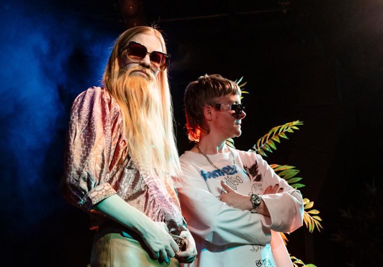 Två dragkings poserar i solglasögon i strålkastarljuset på en scen.