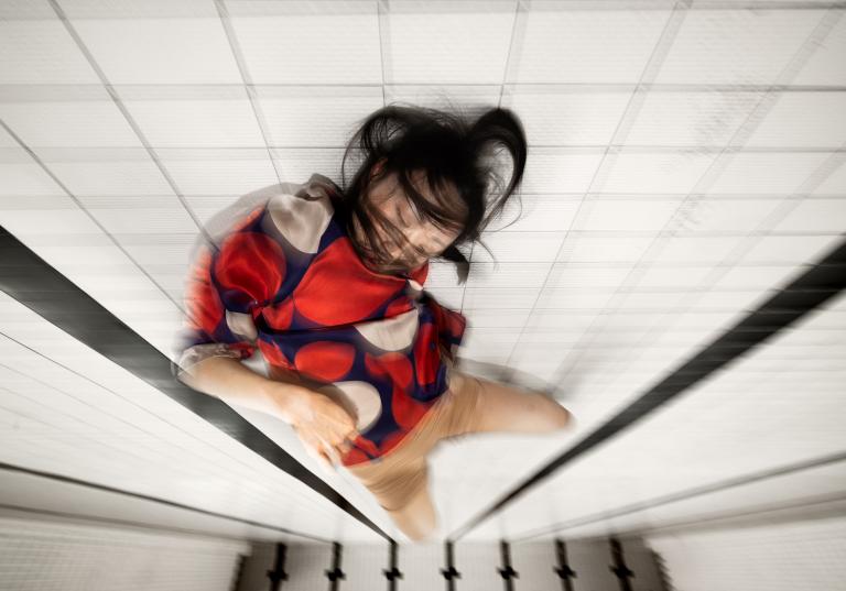 Dansaren Jing Yi Wang i röd och blå kort klänning rör sig på botten av en tömd simbassäng med vitt kakel och långa svarta linjer. Hon blundar och hennes svarta hår sveper runt hennes ansikte.