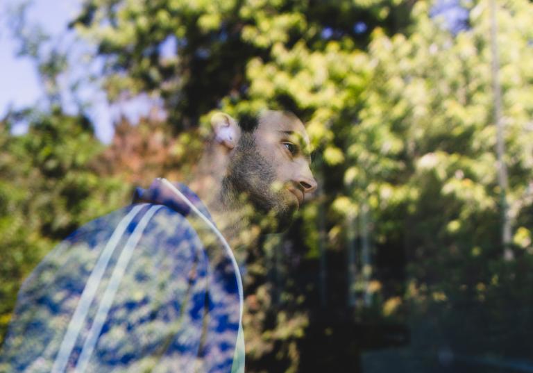 Dansaren Facundo står vid ett stort fönster. Han är klädd i en blå träningsjacka med vita ränder. Han har kort mörkt hår och kort skägg. Facundo täcks nästan helt av reflektionen i fönstret av stora grönskande träd.