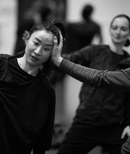 Svart-vit bild på två dansare under repetitioner i studion. Bakom dem skymtas en koreograf. 