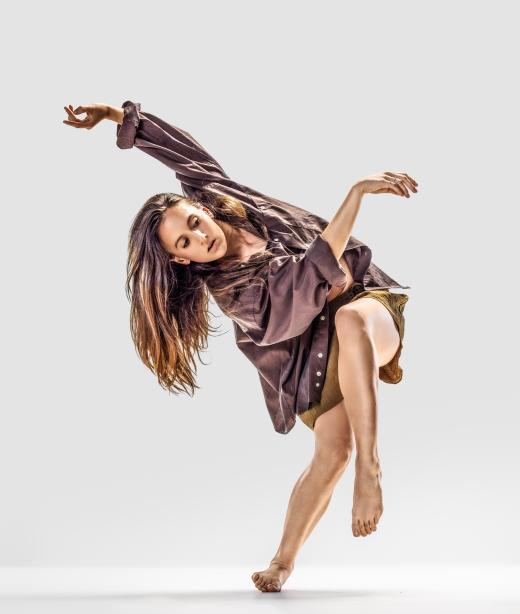 Skånes Dansteaters dansare Hanna Nussbaumer klädd i lila stor skjorta och mörkgula shorts blanserar på ett ben på insidan av sin fot med andra benet böjt framför sig och ena armen sträckt över henne.