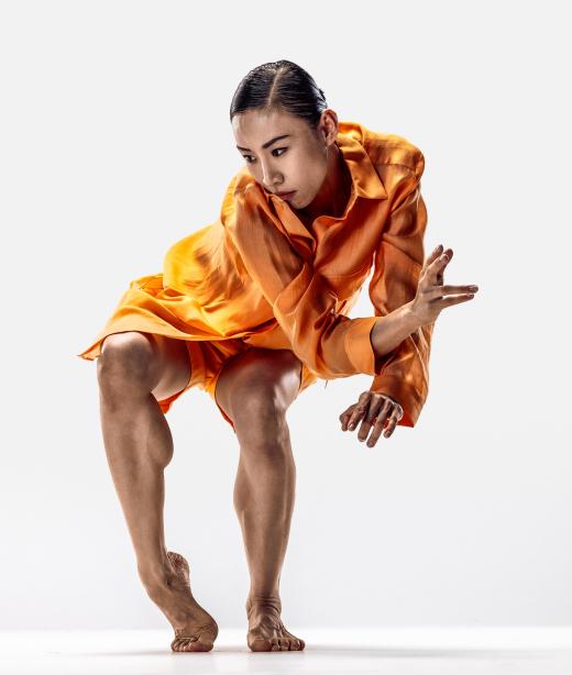 Dansaren Jing Yi Wang klädd i orange shorts och skjorta med svart uppsatt hår dansar med böjda knän och sträcker ut en hand.